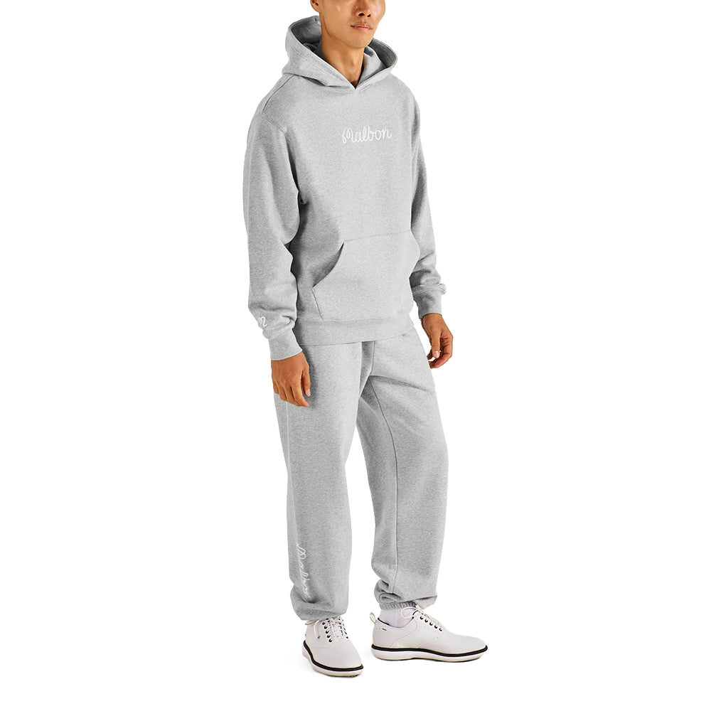 A man modeling a light gray two-piece tracksuit consisting of a pullover hoodie and matching jogger pants, with white sneakers, against a plain white background.