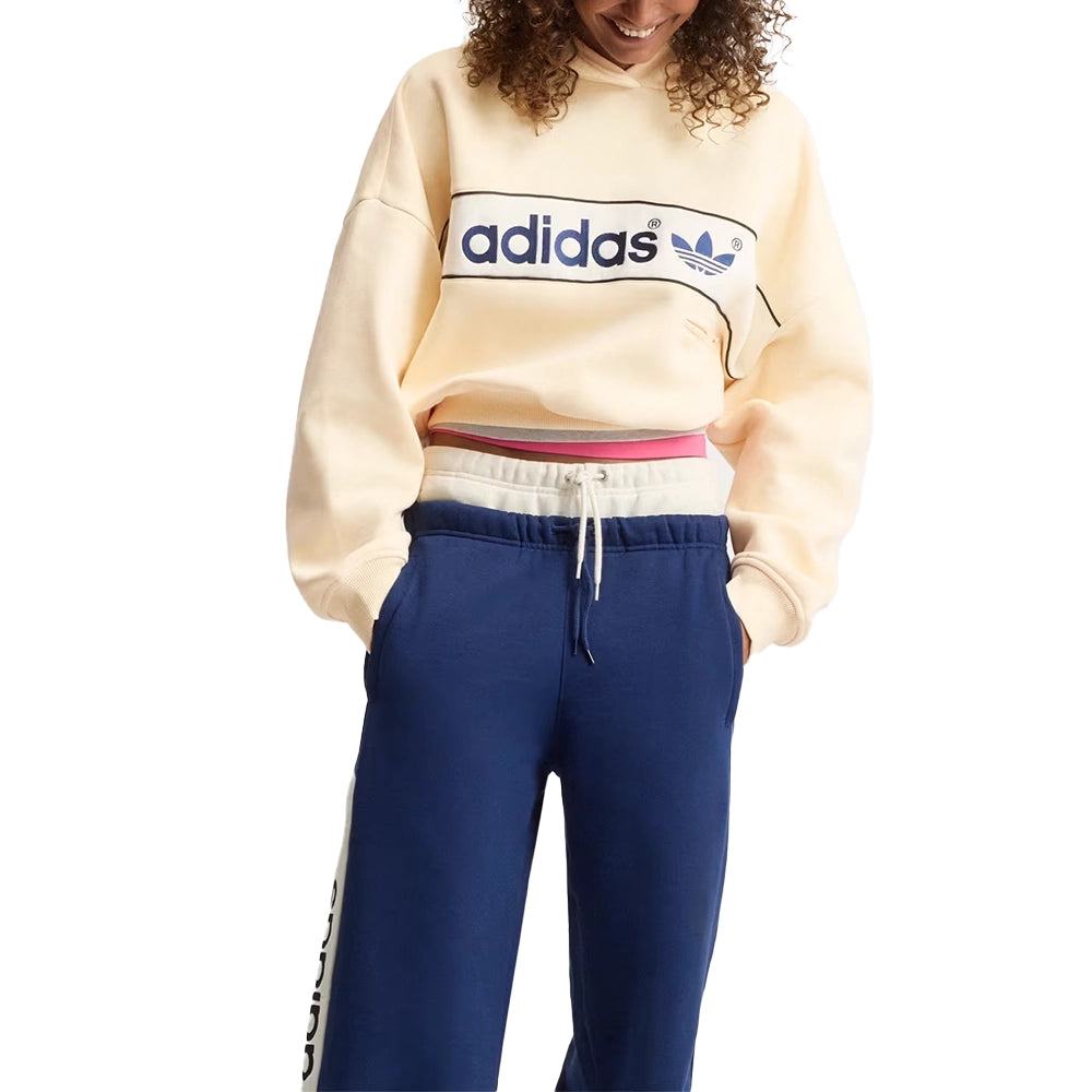 Model wearing the adidas women's adiSignature NY hoodie in Warm Vanilla with navy track pants, front view.
